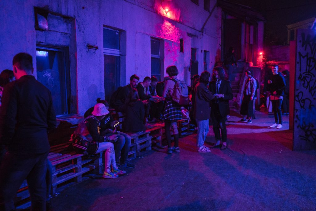 Neon lighting shining on a group of young people in trendy clothes standing and sitting on benches in an alleyway and casually socializing with each other or on their smartphones, as a visual example of the benefits of waitlists for product launch marketing campaigns