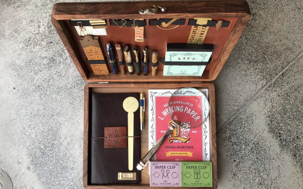 an open wooden box with numerous items inside that are for marketers and copywriters, including paper, magnifying glass, leather notebook, paper clips, pens, and more