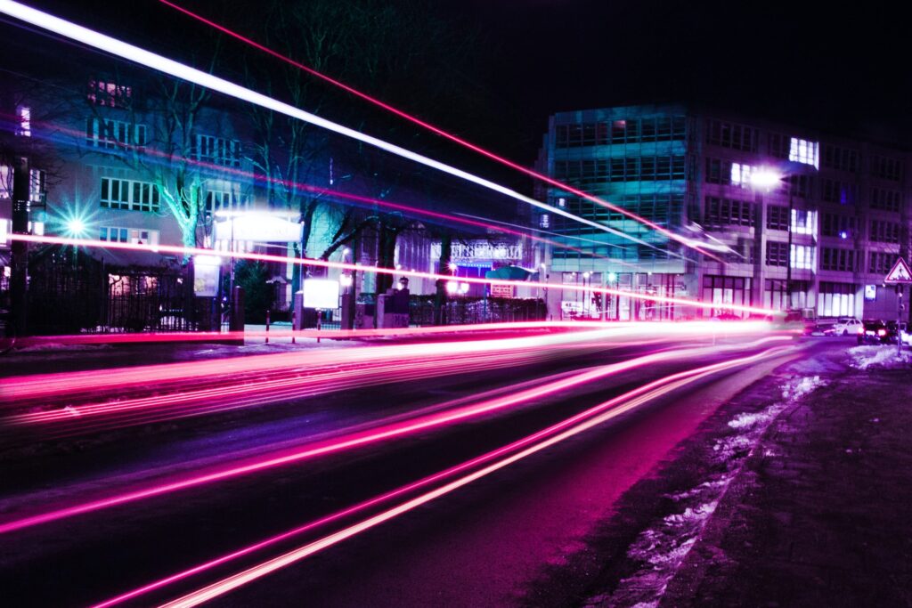 pink and white neon streak lights on a road with buildings in the background lit by purple and green/teal neon lights; the background is black (nighttime)