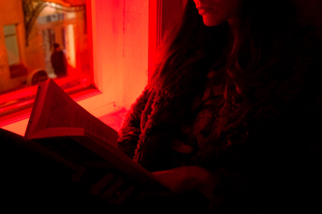 the best books to read in 2021, recommended for your reading list by business and marketing executives: image of someone reading by a window in shadow with red and orange neon light illuminating them through the window