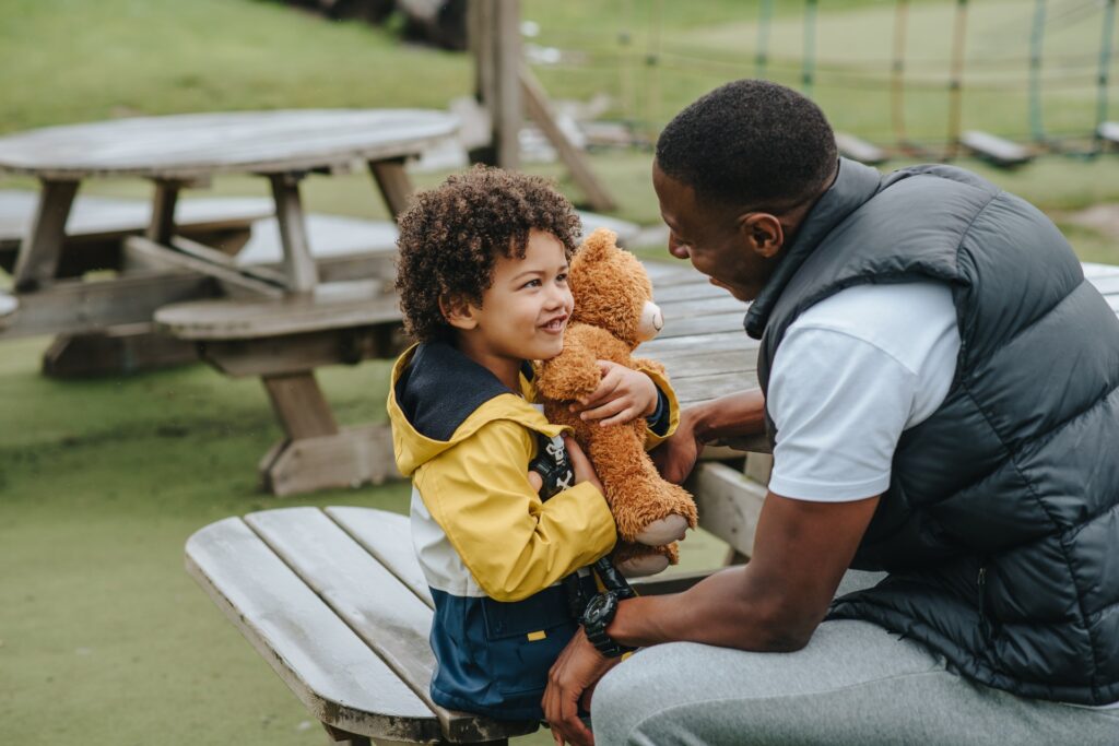 image of a little brown skinned boy holding a teddy bear and looking adoringly up at a sweetly smiling black man, presumably his father, while they sit on picnic table benches outdoors